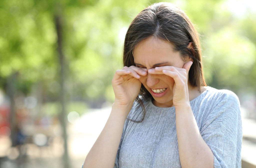 A woman with dry eyes is rubbing her eyes while standing outdoors in a park
