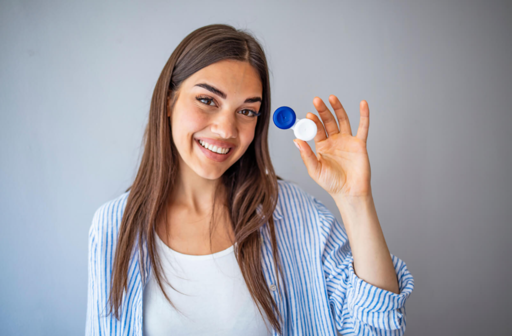 A young woman against a grey background smiling and holding up a contact lens case with her left hand.