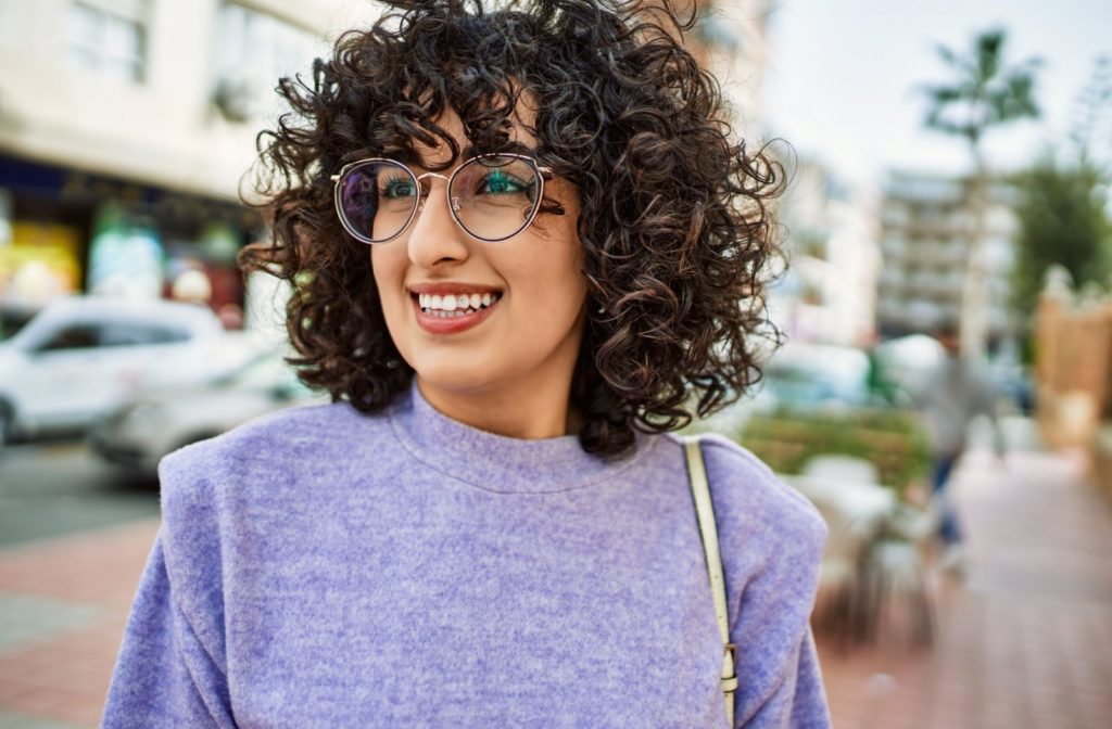 A curly-haired woman wearing stylish glasses and a purple sweater, smiling while standing outside.
