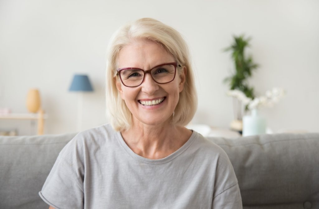 A relaxed older woman wearing glasses sits on a couch and smiles confidently.