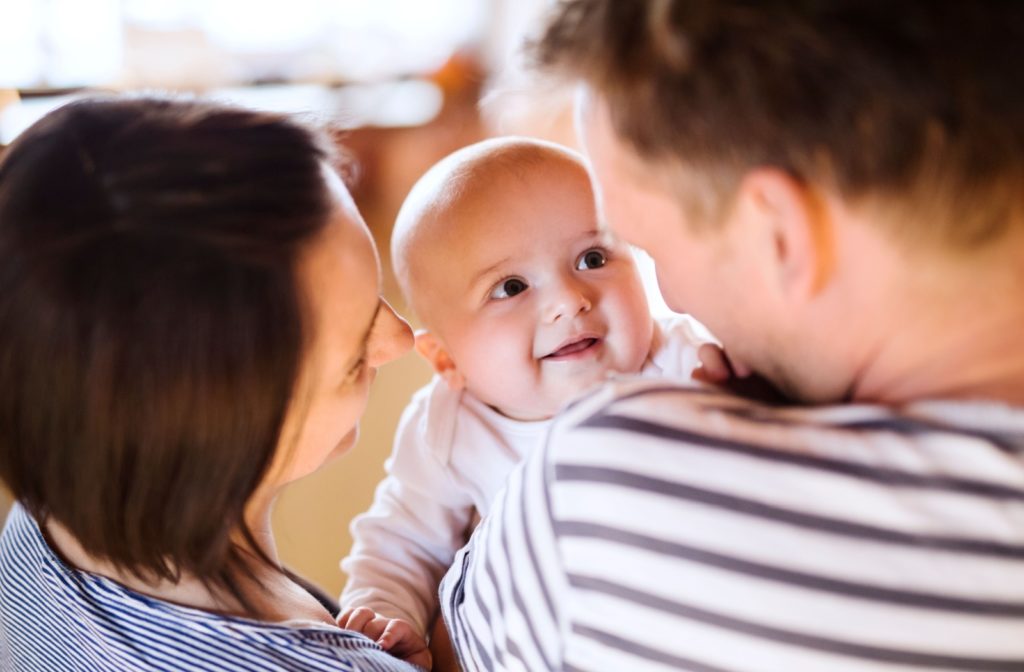 A smiling baby faces the camera, held between their two parents, who have their backs to the camera