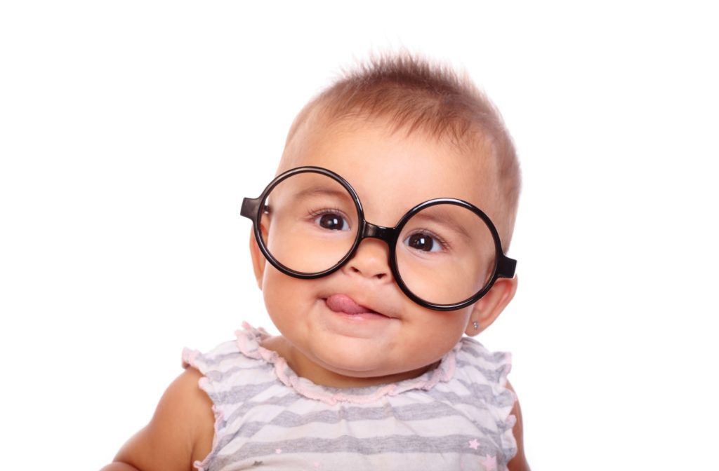 A young baby against a white background with black-rimmed circular glasses looks to the left while sticking out their tongue