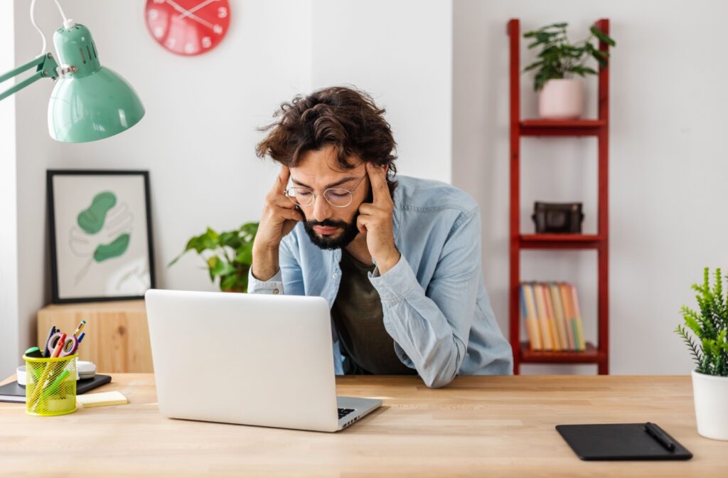Man wearing glasses sitting at a desk, holding his head with both hands and looking at a laptop, experiencing a headache caused by cataracts.