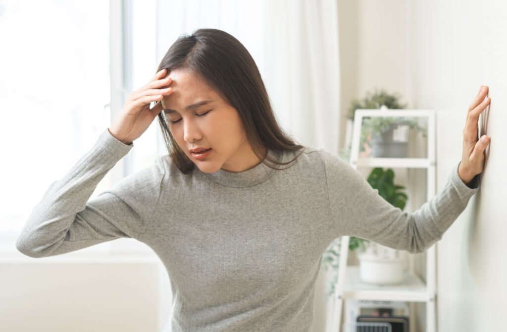 Woman holding her forehead with eyes closed and leaning against a wall, appearing dizzy or lightheaded.