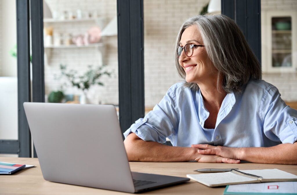 Senior person wearing glasses, smiling while looking out a window and sitting in front of a laptop.