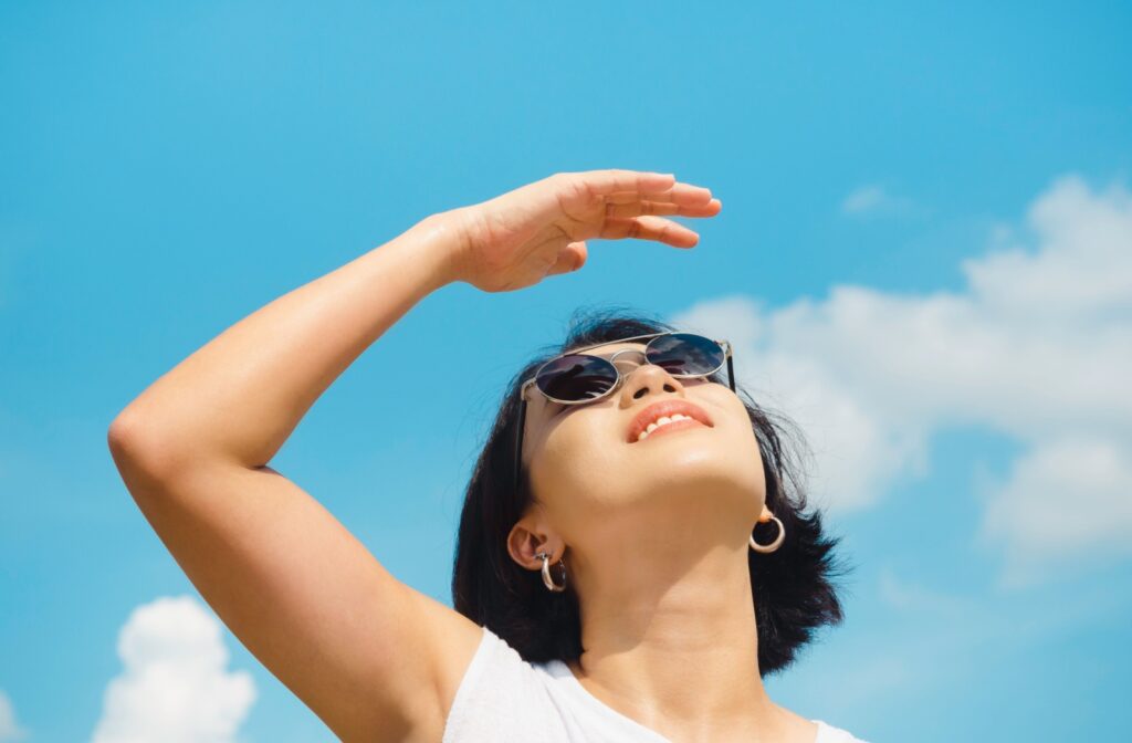 A person wearing sunglasses, looking up at the sun, using their hand to cast a shadow over their face.
